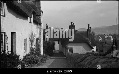 Dunster, West Somerset, Somerset, 1940-1953. A view of the town taken from a small street on a hill. There is a whitewashed thatched cottage on the left hand side in the foreground, and the street slopes downwards to the midground. In the background the roofs of houses below can be seen. Stock Photo