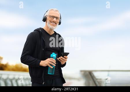 Senior man using phone while have outdoor workout, drinking water Stock Photo