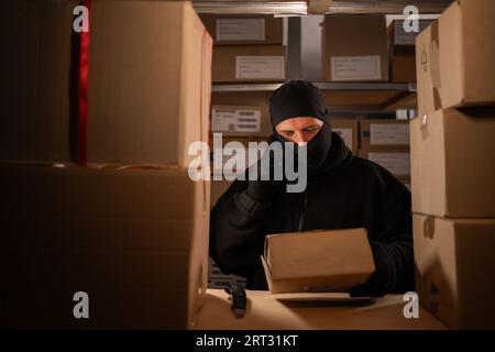 Thief stealing a box in a warehouse calls on a mobile phone at night. Concept of problems with the theft of goods and parcels at the post office. Stock Photo