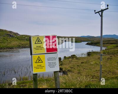 Yellow and red signs at a lochan (small lake) on Great Bernera in the Outer Hebrides, Scotland, UK warning anglers to cast with care near overhead pow Stock Photo