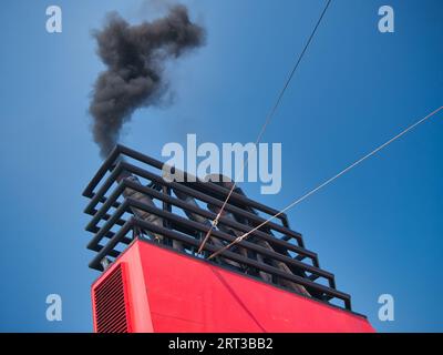 Thick black smoke leaves the exhaust on a ship with surrounding black superstructure. Taken on a sunny day with a blue sky. Stock Photo