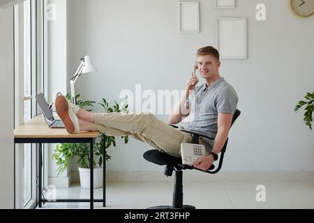 Smiling man sitting at desk with legs on table and talking on phone Stock Photo