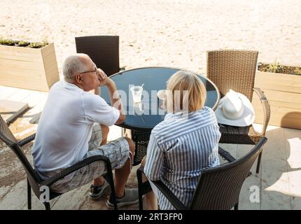 An elderly couple sits at a small table in an outdoor cafe near the sea, drinking coffee and chatting. Stock Photo