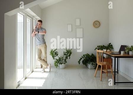 Businessman standing at home office window, checking time and on wristwatch and talking on phone Stock Photo