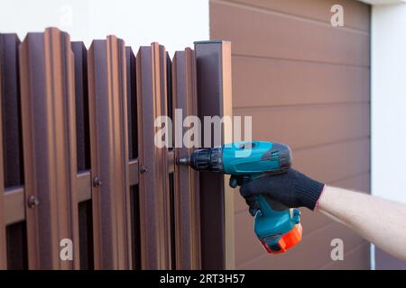 Screwing roofing screws into a metal picket fence by using cordless electric screwdriver. Fencing, installing the fence. Close up. Stock Photo