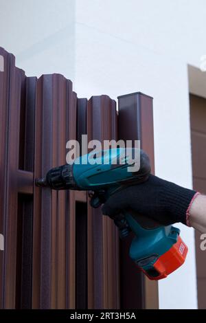 Screwing roofing screws into a metal picket fence by using cordless electric screwdriver. Fencing, installing the fence. Construction work. Close up. Stock Photo