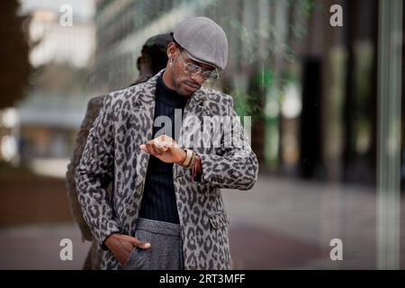Handsome black man in stylish clothing looking at wristwatch while standing near glass wall in city. He is wearing trendy cap, coat and glasses. Stock Photo