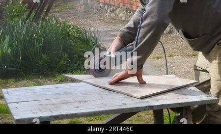 cutting finishing tiles using a grinder in an outdoor tiler's workshop, a man cuts the corner of a ceramic tile with an angle saw Stock Photo