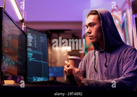 Asian hacker drinking coffee and doing ddos attack on website. Criminal breaking into network system on computer screen while holding beverage to go in abandoned warehouse Stock Photo