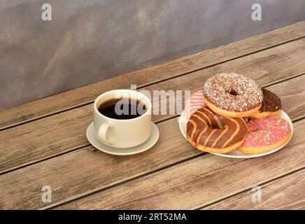 A cup of hot black coffee on a saucer and a plate with a bunch of fresh donuts in multi-colored glaze on a wooden table against a gray wall. Close-up. Stock Photo