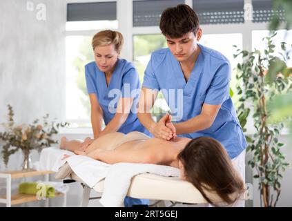 Two female and male masseuses perform stroking and rubbing of muscles of back and shoulders to girl Stock Photo