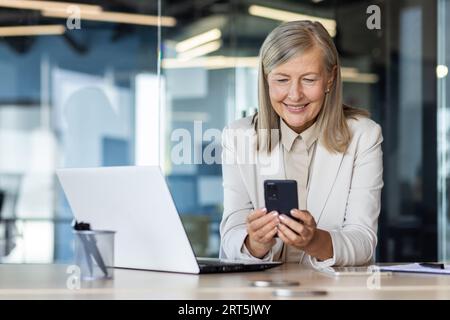 Senior mature experienced woman at workplace inside office, business woman happy smiling using phone sitting with laptop, gray haired woman boss using app on smartphone typing message. Stock Photo
