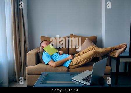 Young adult caucasian man felt asleep on a couch dropped book on face. Tired man sleeps on sofa Stock Photo