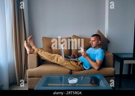 Young adult caucasian man chats on phone and works on laptop lying on sofa. Thoughtful man texting lying on couch Stock Photo
