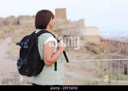Active middle aged woman with backpack relaxes, looks at beautiful view after hiking up. Healthy lifestyle in retirement. Adventure seeks woman treks enjoys freedom. Stock Photo
