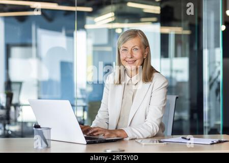 Portrait of senior mature gray haired businesswoman inside office, successful adult woman smiling and looking at camera, boss working with laptop in business suit looking at camera smiling. Stock Photo