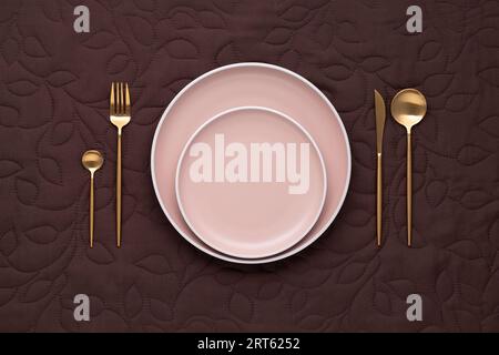 Festive table setting. Golden fork, knife and spoon. Empty ceramic plate with gold cutlery. Dark brown tablecloth. Top view. Dining in luxury restaura Stock Photo