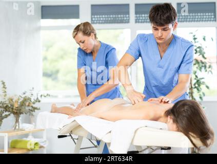 Young masseur doing back massage to woman in tandem with female colleague Stock Photo