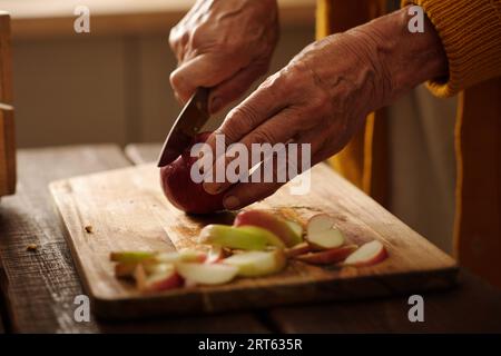 Hands of grandmother with sharp knife slicing fresh ripe apples on wooden board while making preparation of fruits for winter Stock Photo