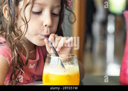 Close-up portrait of a cute little white Caucasian girl drinking a glass of fresh orange juice with a straw Stock Photo
