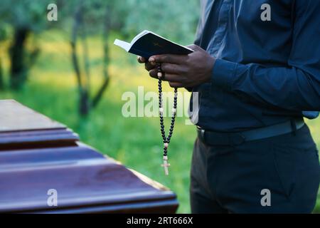 Close-up of African American man in priest attire holding rosary beads and open Holy Bible while standing in front of coffin during burial service Stock Photo