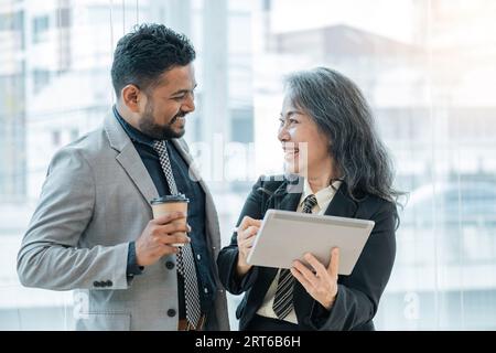 Diverse friendly coworkers colleague indian male manager and asian female employee talking in office using tablet device together discussing project Stock Photo