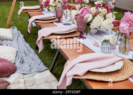 Aesthetics pink cozy picnic, served table with pink flowers bouquets, pillows, candles decor shells in the garden. Stock Photo