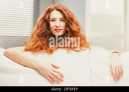 Peeking from behind plush sofa cushions, a joyous woman beams amid her fiery long, billowy red hair. Lounging gracefully on her white couch, her slend Stock Photo