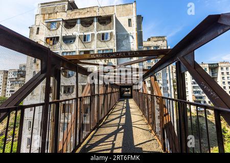 A walkway of the Tbilisi Skybridge residential complex (Nutsubidze Skybridge) on Shalva Nutsubidze Street in Tbilisi, Georgia, leads between rusty steel lattice girders towards a tower block. Side fences protect the walkway from the wind. The girders cast shadows on the floor. The tower shows exposed concrete and horseshoe-shaped window openings, with apartment blocks on the hillside to the right. The complex connects the Saburtalo and Nutsubidze neighbourhoods on the hillside. It lies around 11 kilometres from the historic city centre. Architects Otar Kalandarishvili and Gaioz Potskhishvili d Stock Photo