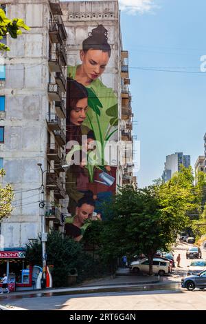 A mural spanning many storeys on the gable wall of a prefabricated block near the Tbilisi Skybridge residential complex in Tbilisi, Georgia, shows five female figures with dark hair around a large green plant in a pot. One of the figures waters the plant with a watering can. At the top, a young woman with her hair in a bun and a green top looks down. A shop with cash machines occupies the ground floor. Cars drive along the street. A man walks along the pavement. Trees line the street on the right. The Tbilisi Skybridge complex links three high-rise blocks and the Saburtalo and Nutsubidze neigh Stock Photo