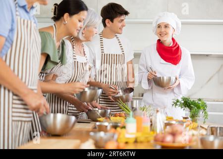 Female chef giving instructions to students standing around table in cooking class Stock Photo