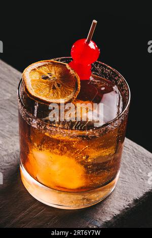 High angle of glass filled with alcoholic old fashioned whiskey and ice cubes garnished with dried lemon slice and cherries placed on table Stock Photo