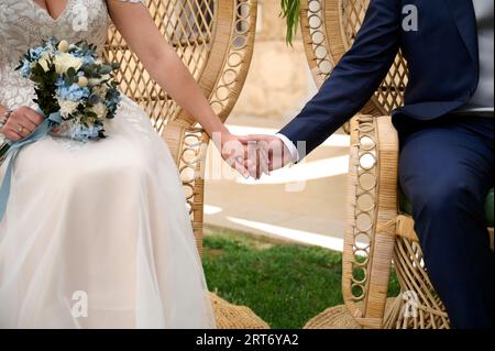 Unrecognizable bride and groom holding hands while sitting on wicker chairs against blurred background of nature Stock Photo