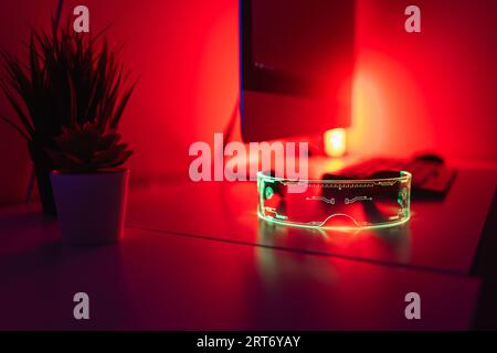 Wooden workplace desk mini potted green plants AR glasses computer monitor keyboard mouse in bright glowing red neon light in blurred corner of room Stock Photo