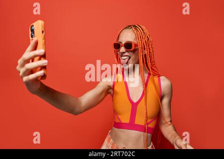 Young stylish woman with Afro braids in sunglasses and colorful top taking selfie on cellphone against red background Stock Photo
