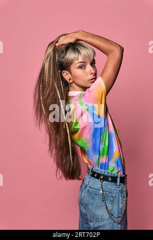 Young female hipster wearing colorful t shirt and jeans touching long hair while looking over shoulder against pink background Stock Photo