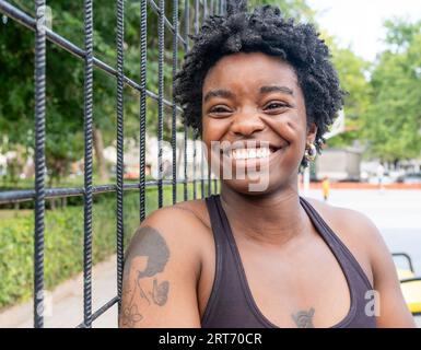 Happy black tattooed sportswoman with short Afro hair smiling widely while standing near metal fence on sports ground during fitness workout Stock Photo