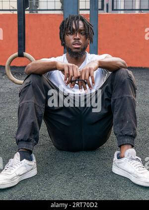African American male with dreadlocks in activewear resting on floor in sports ground and looking at camera Stock Photo