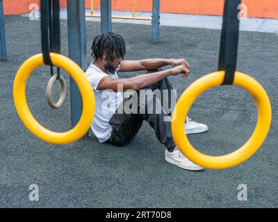 Full body side view of young African American man with dreadlocks resting on sports ground leaning on metal bar during break from training Stock Photo