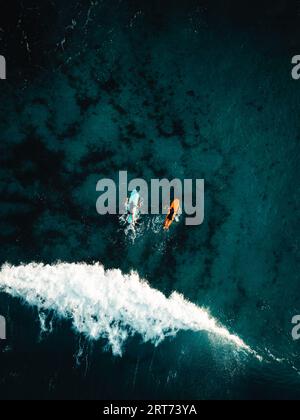 Two surfers floating on surfboard in tropical ocean with big and powerful wave behind them. Aerial top view of surfers in crystal blue ocean Stock Photo