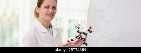 Female scientist standing near whiteboard, pointing to molecular model in her hand. Stock Photo