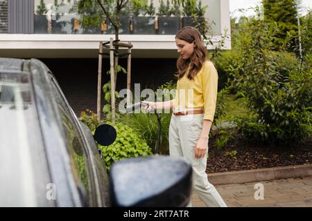 Side view of beautiful woman plugging charger in her electric car. Progressive woman charging her electric car on the street. Stock Photo