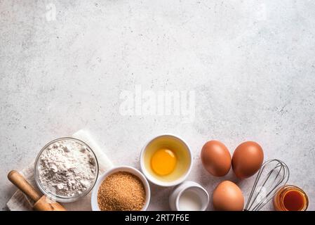Wheat flour, eggs, rolling pin and whisk on white concrete background, top view, copy space. Ingredients for bakery, baking or cooking concept. Stock Photo