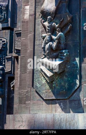 A bronze relief above the base of a pillar of the Chronicle of Georgia (Georgian საქართველოს მატიანე, Sakartvelos Matiane) on Mount Kenisi above the Tbilisi Sea, a reservoir, in the Nadzaladevi district of Tbilisi, Georgia, shows a standing bearded figure. It raises one arm beneath a billowing cloth and holds a scroll. In front of it, a man with long hair and a beard possibly sits on a stack of books. A large book forms the base of the scene. Verdigris covers the bronze. On the left, plaques bear Georgian inscriptions. Sculptor Zurab Tsereteli began the complex in 1985. It consists of 16 pilla Stock Photo