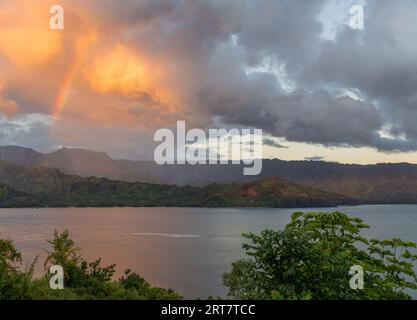 Sunrise illuminates storm clouds and rainbow over Hanalei mountains from Princeville overlook of bay Stock Photo