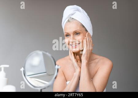 Beautiful mature woman with towel on head looking at mirror, using new brand anti-aging cosmetics, home interior Stock Photo