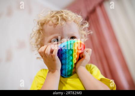 girl covered her face with a multi-colored round pop it. Blond girl playing with pop it sensory toy. fashionable silicone game for stressed children Stock Photo