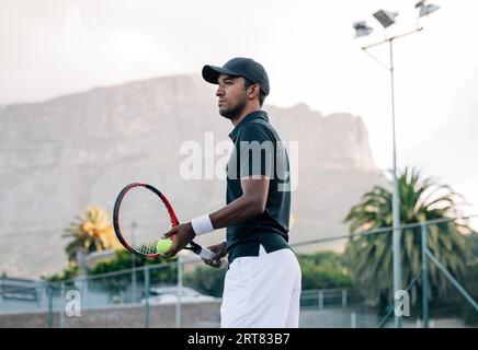 Fit man in sportswear and cap with a racket standing on tennis court. Tennis player looking away while standing on a hard court. Stock Photo