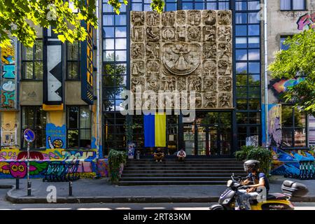 A large Soviet-era relief dominates the glass facade of Fabrika (Georgian ფაბრიკა, "factory") on Egnate Ninoshvili Street in the Chugureti district of Tbilisi, Georgia. In the centre a female figure spreads her arms inside a circle. Relief panels with figures, animals and plants surround her. A Ukrainian flag hangs above the entrance. Two men sit on the front steps. On the left a vertical sign bears the Fabrika lettering. The side walls are covered in graffiti. In the foreground a man rides past on a motor scooter. Fabrika occupies a Soviet-era sewing factory that made women's clothing. Since  Stock Photo