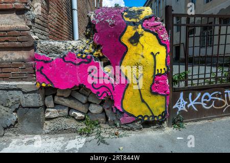 A painted fragment of wall stands beside a pavement in the Chugureti district of Tbilisi, Georgia. On a pink background it shows a stylised yellow figure with a black outline. The painted layer has broken off at the edges. Loose rubble stones lie behind it. A brick wall adjoins it on the left. A metal gate with a white tag stands on the right. Stock Photo